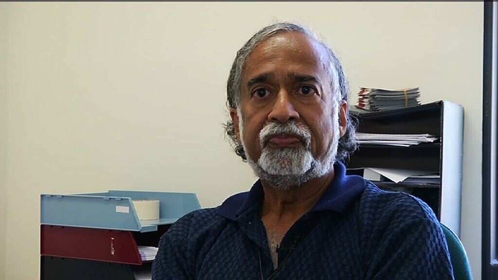 A man in blue jumper with white beard in front of filing cabinets.
