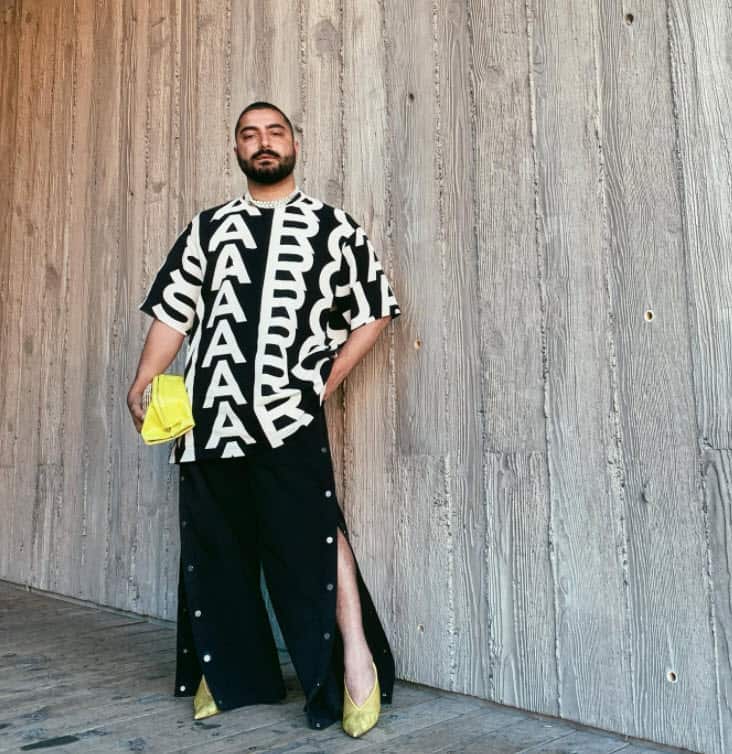 A non-binary person with a beard wears a long flowing shirt, skirt and high heels.