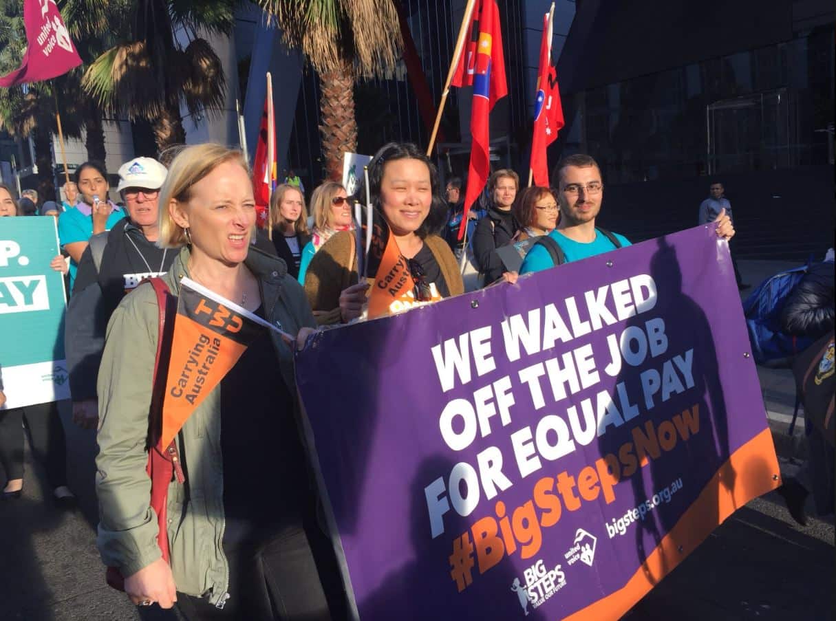 People are walking in a protest, holding a poster that reads 'We walked off the job for equal pay,' with others carrying posters and flags behind them.