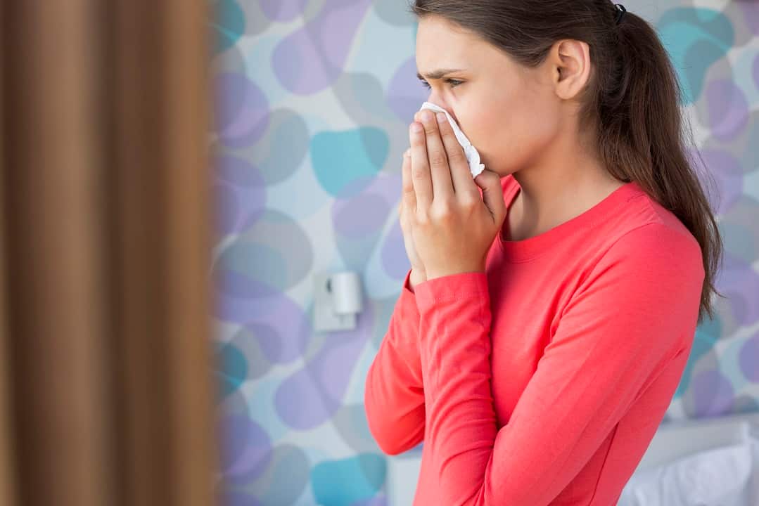 A woman with long hair blowing her nose with a tissue while standing.