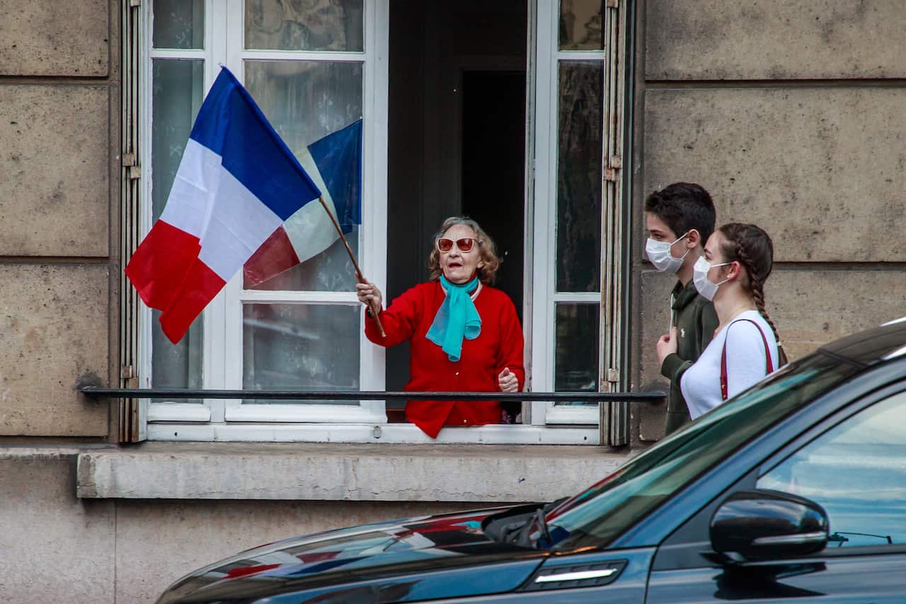 A couple wearing protectives face masks pass by a building where a woman holds a French flags. 