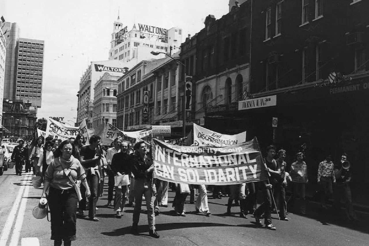 A black and white photo of people marching down Sydney's Oxford Street with gay rights banners. Two people at the front hold a large banner reading: "International gay solidarity".