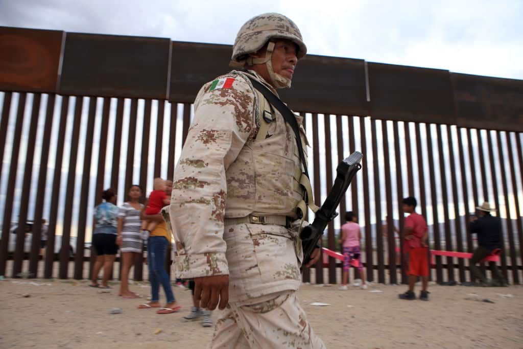 A mexican soldier walks in front of American and Mexican families playing on the seesaw.