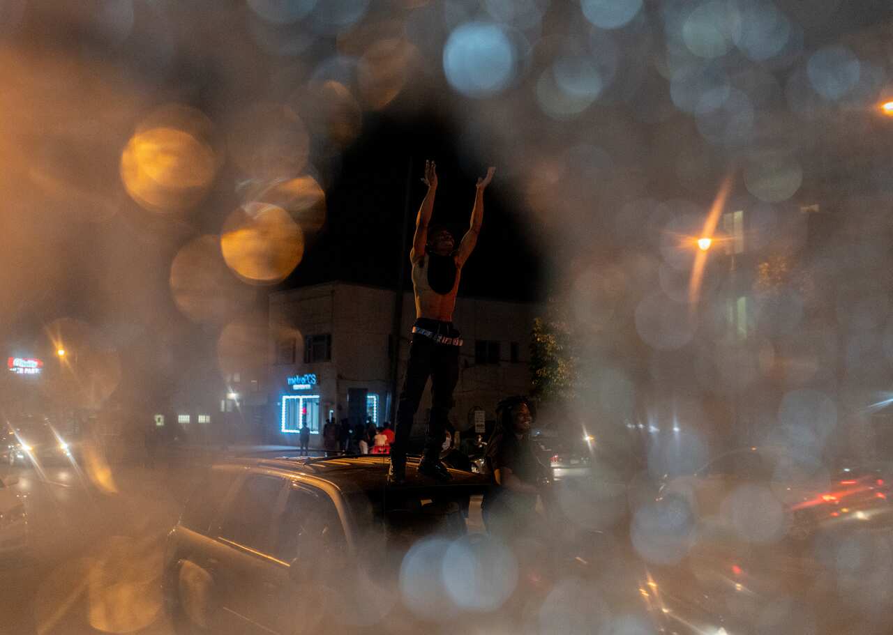A man, seen through the broken glass of a bus stop, stands on a car and claps during a protest near the Third Police Precinct.