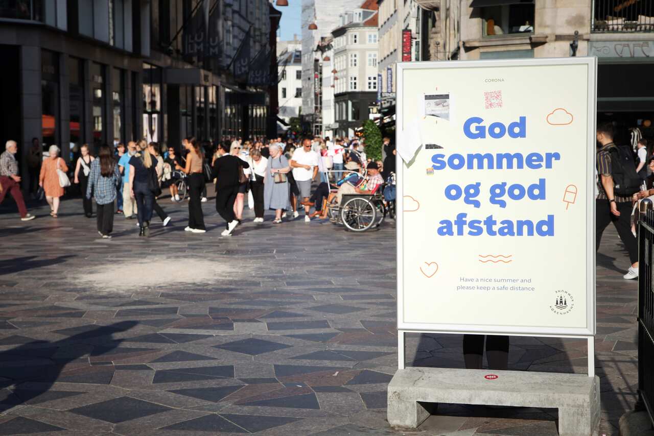Passers-by walk along Strget, Copenhagen's central shopping street. In front of them is a faded sign that reads "God sommer og god afstand" (Good summer and good distance).