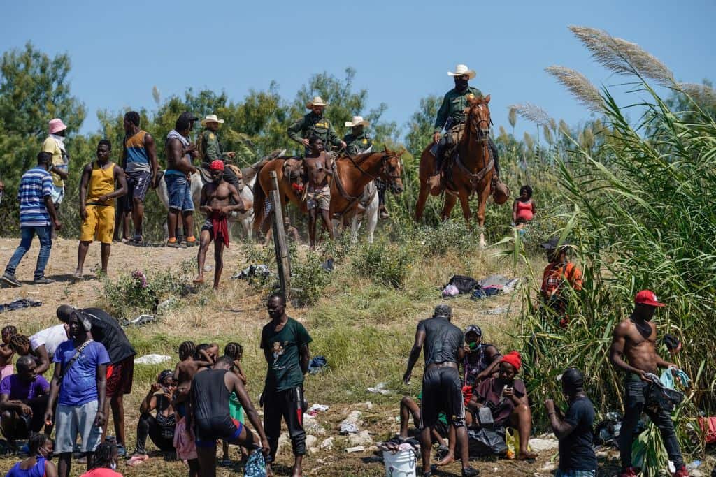 US Border Patrol agents on horseback look on as Haitian migrants sit on the river bank near an encampment on the banks of the Rio Grande, Texas. 