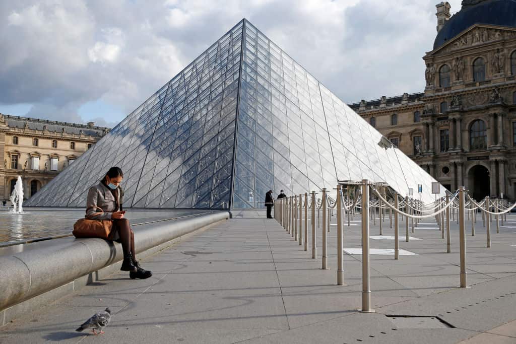 A woman wearing a protective face mask seats in front of the empty courtyard of the Louvre museum in Paris, France.