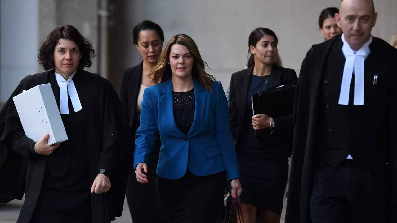 A woman in a blue jacket walking, flanked by lawyers.