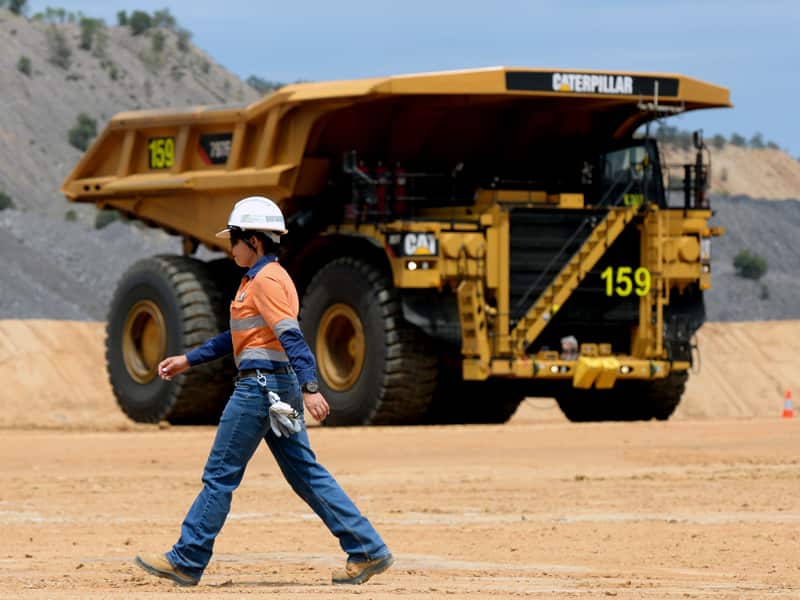 A woman in orange and blue hi-vis shirt and long blue trousers walking on packed earth in front of a very large yellow truck