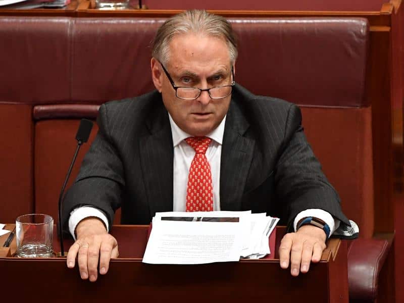 A man in glasses and a suit sits at a lectern and looks into the camera.