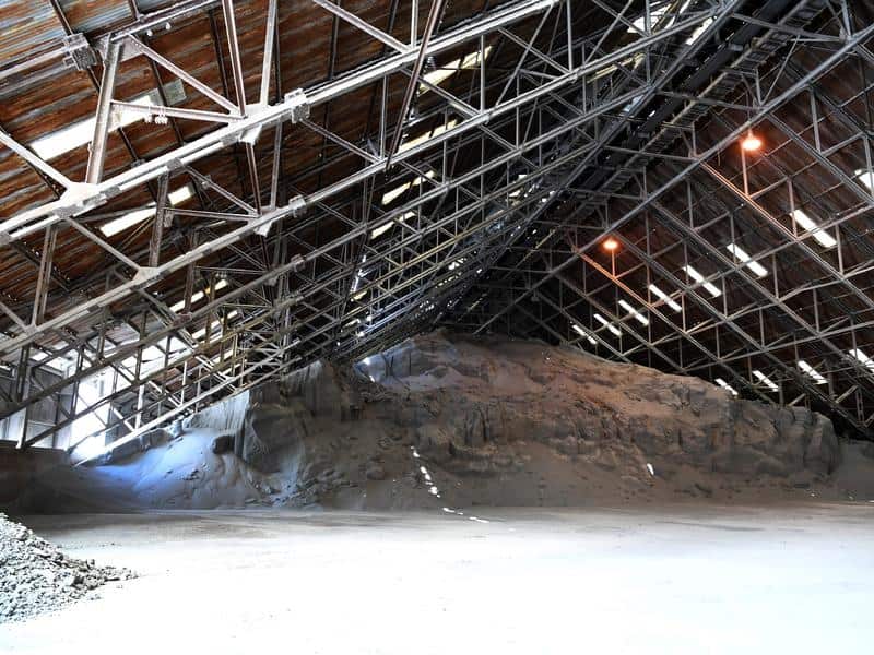 A vast pile of grey industrial material is stored inside a large warehouse featuring a complex network of exposed steel roof trusses and rows of overhead lights.