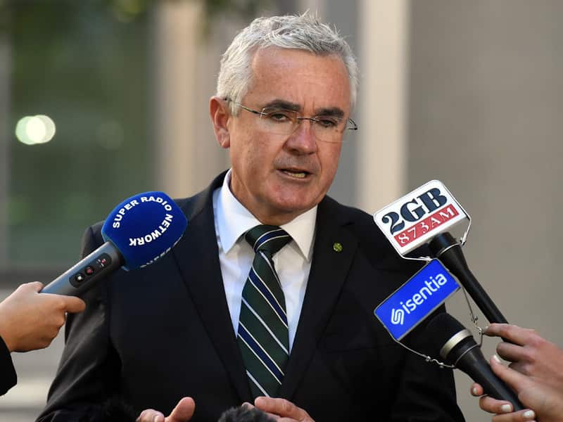A man in a suit and tie speaking. People are holding microphones in front of him