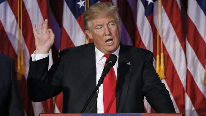 Donald Trump standing at a lectern. He is wearing a dark suit and red tie and holding up his right hand. There is a US flag in the background