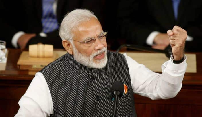 Narendra Modi sitting behind a microphone and holding up his left fist.