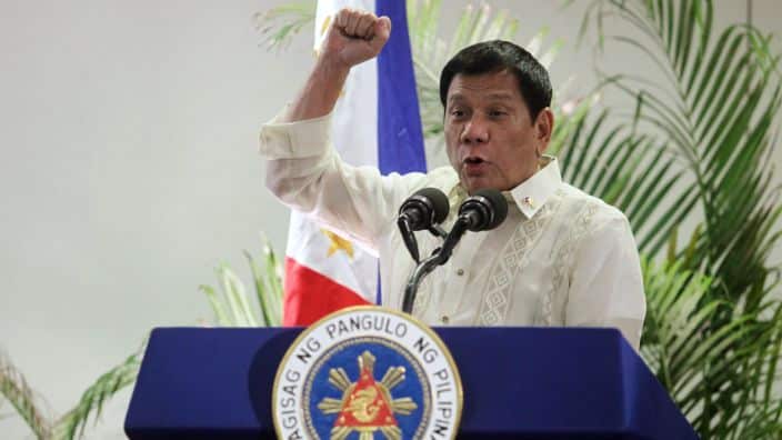 Man wearing a white shirt standing behind a lectern and talking. His right fist is raised.