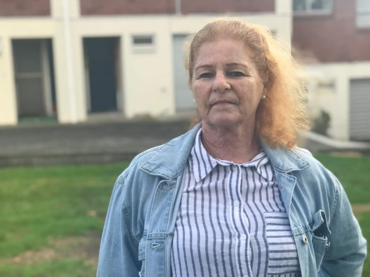 An elderly woman wearing a denim jacket over a white shirt with blue stripes stands in front of townhouses.