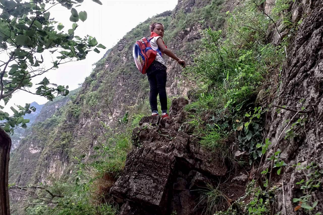 A child wearing a backpack stands on a rock, a sheer cliff behind her.