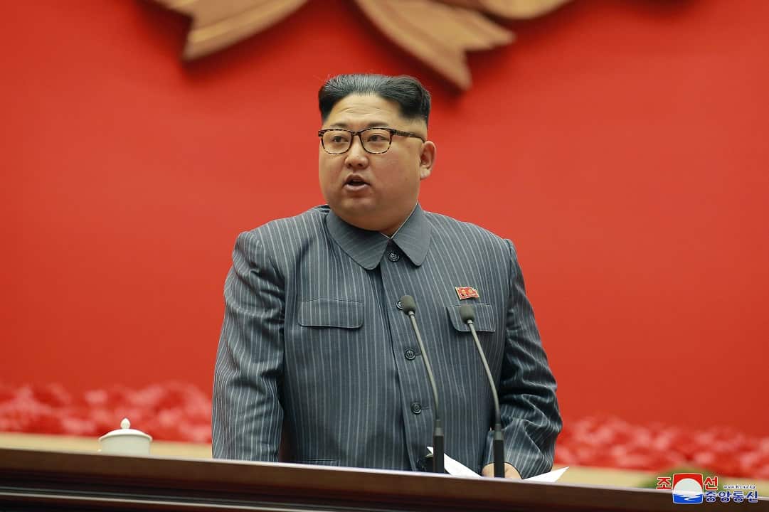 North Korean leader Kim Jong-un wearing a grey striped suit and standing in front of a red backdrop