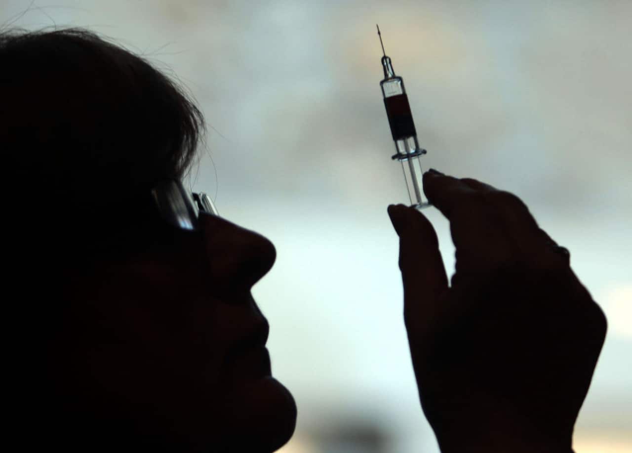 A silhouetted profile of a person wearing glasses looking upward at a syringe held in their hand against a bright, blurred background.
