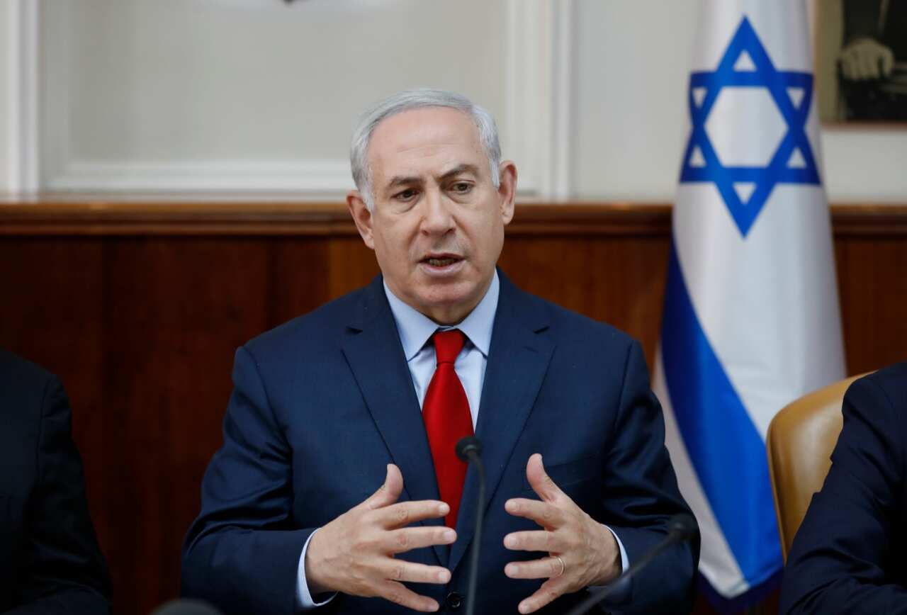 Israeli Prime Minister Benjamin Netanyahu speaks into a microphone. And Israeli flag is hangs limply behind him