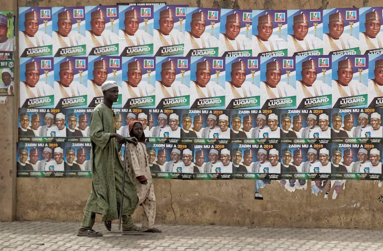 A child leads a blind man as they walk past a wall of campaign posters in Kano, in northern Nigeria, Saturday, Feb. 16, 2019. 