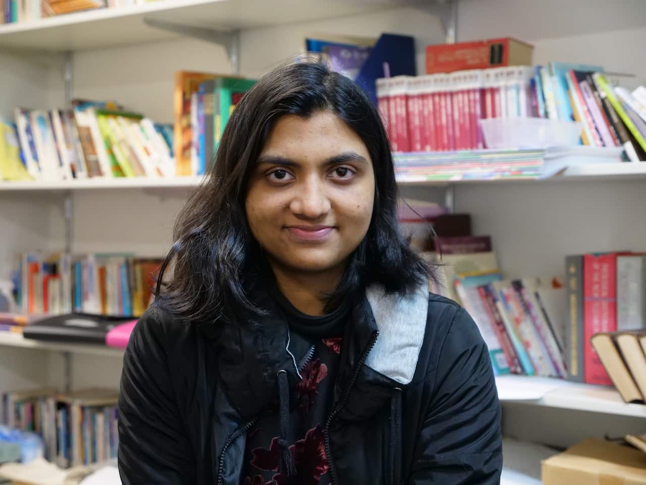 A young woman smiling at the camera. She's sitting in front of a bookshelf