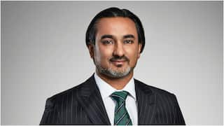 A studio headshot of a man with dark, slicked-back hair and a trimmed beard, looking forward and smiling faintly while wearing a pinstripe suit, a white shirt, and a green striped tie against a plain gray background.