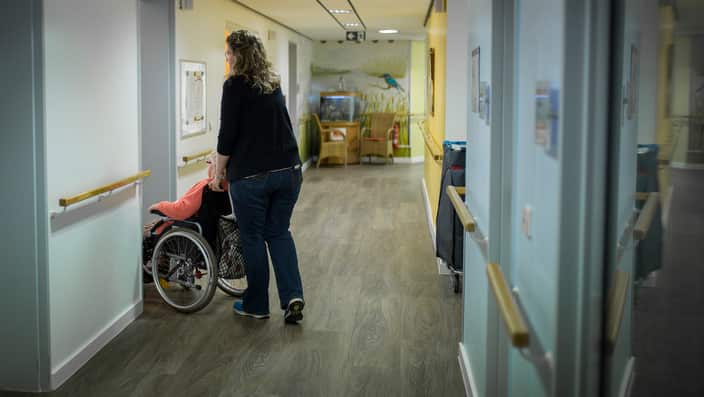 A caregiver pushes an older woman in a wheelchair through a corridor  in a care facility 