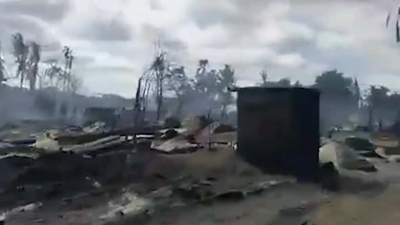 A hazy image of a lone structure standing amidst tall trees and rubble as smoke billows behind.