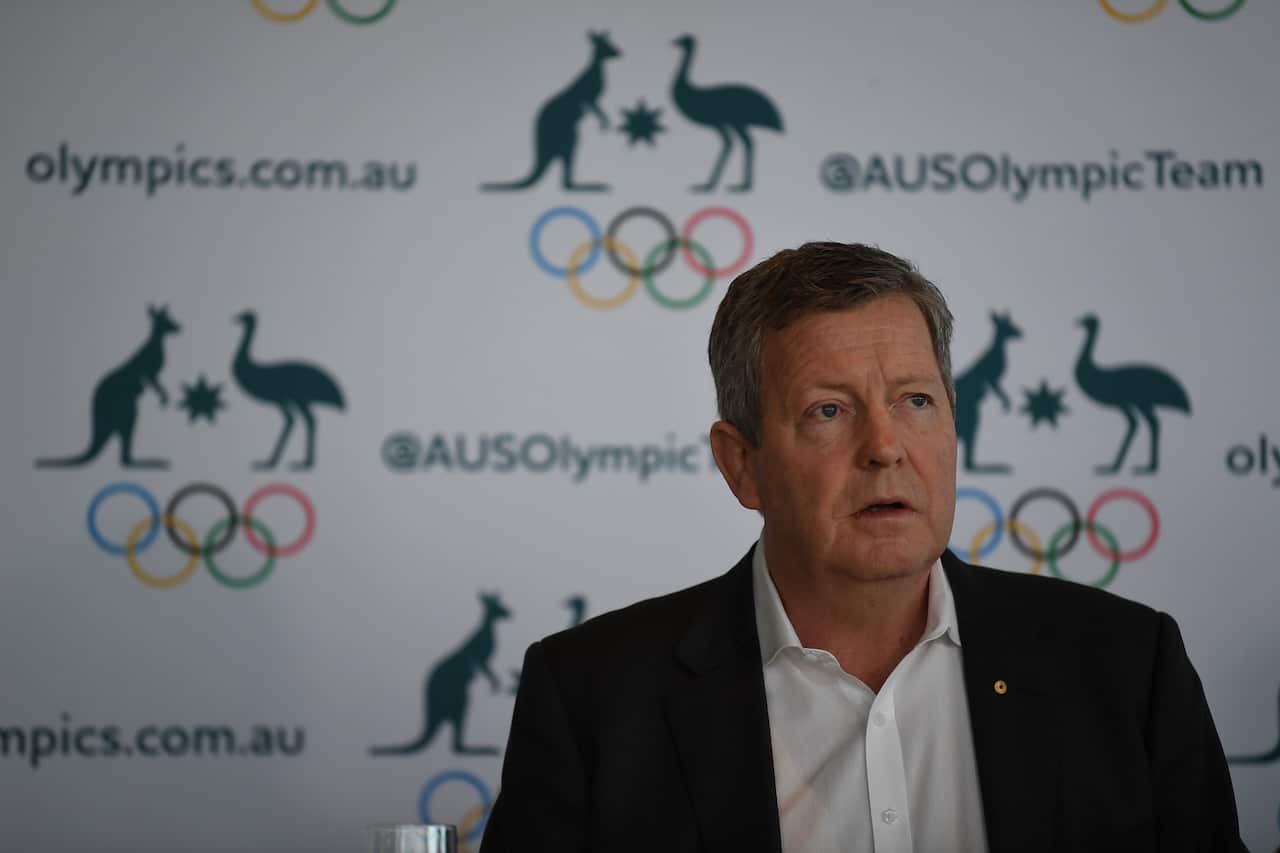 A man in a black suit stands in front of a board with the Olympics logo and Australian coat of arms. 