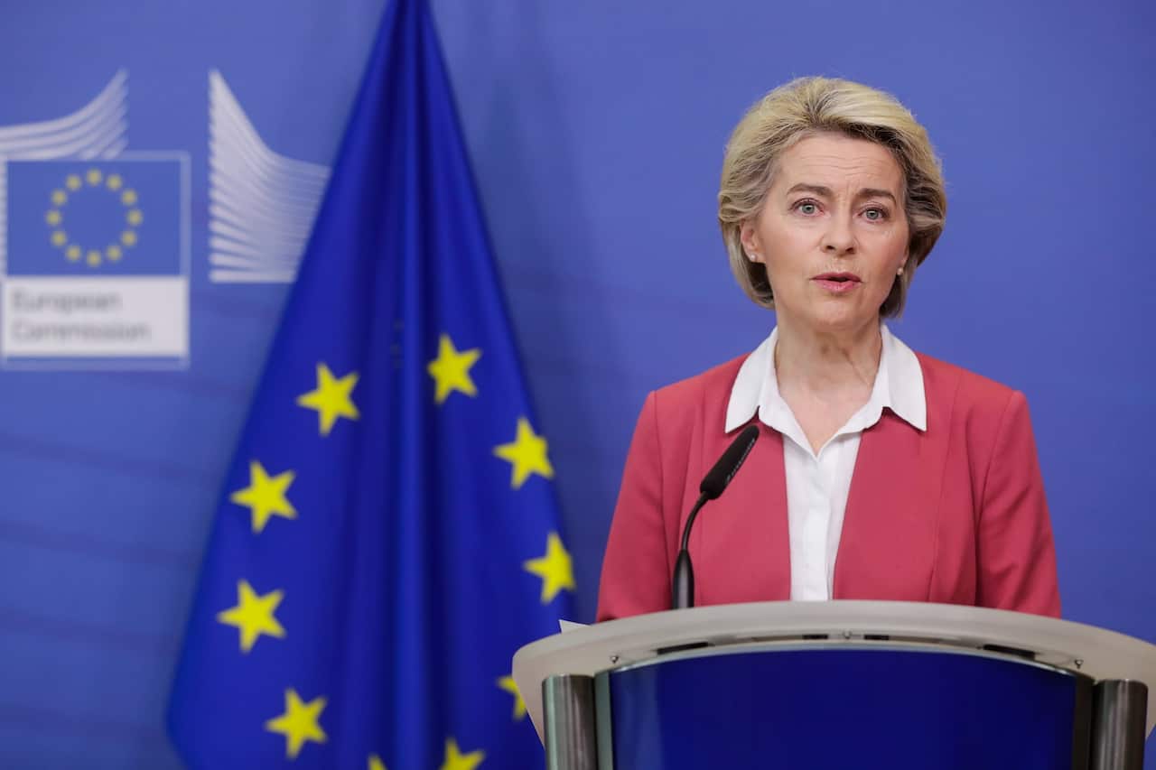 Ursula Von Der Leyen, wearing a red jacket and white shirt, stands talking at a lectern. There is a blue flag with yellow stars behind her right shoulder