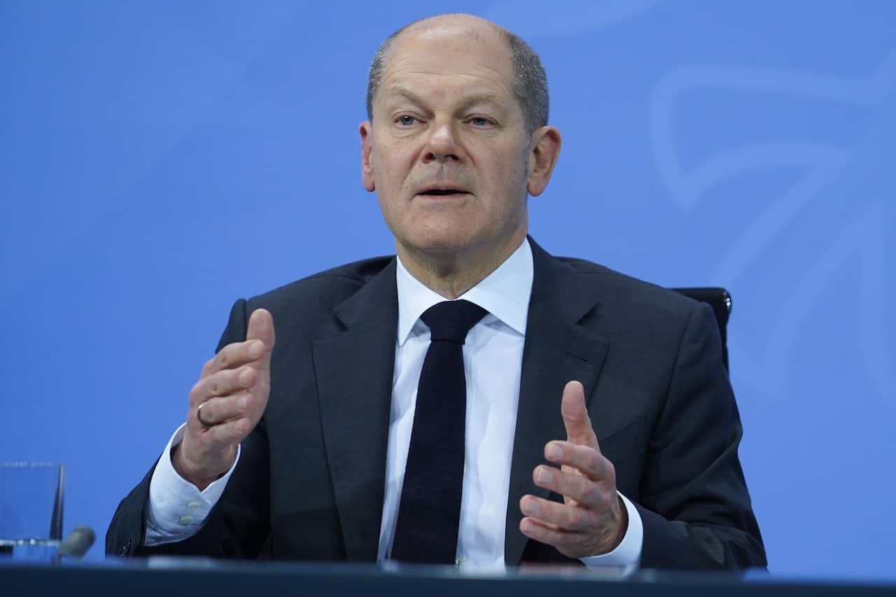 A man wearing a suit and tie speaks while seated at a desk.