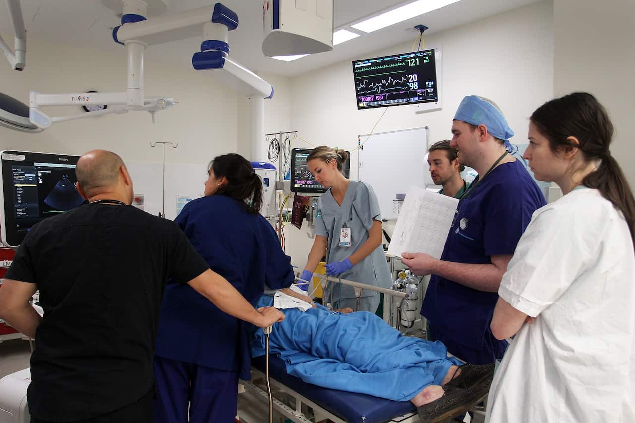 A crowd of doctors and nurses wearing scrubs in various shades of blue crowds around a person lying on a bed with a blue blanket in the emergency department.