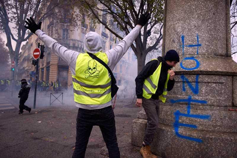 A yellow vest with the words 'block everything' on his back gestures toward riot police.