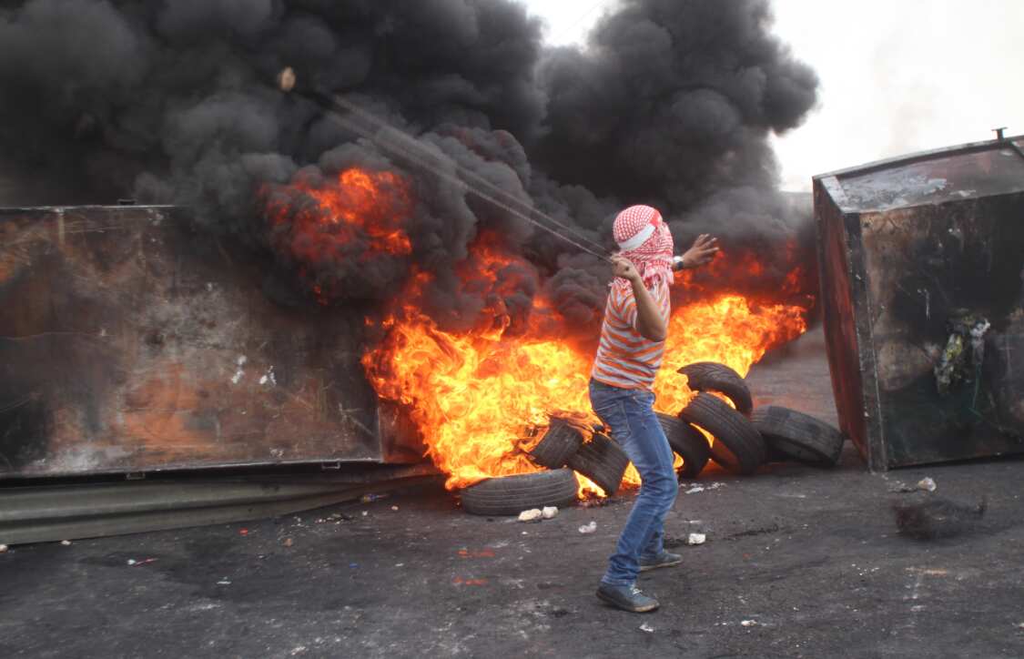 An Palestinian uses a slingshot to throw a rock, tyres burn in the background. 