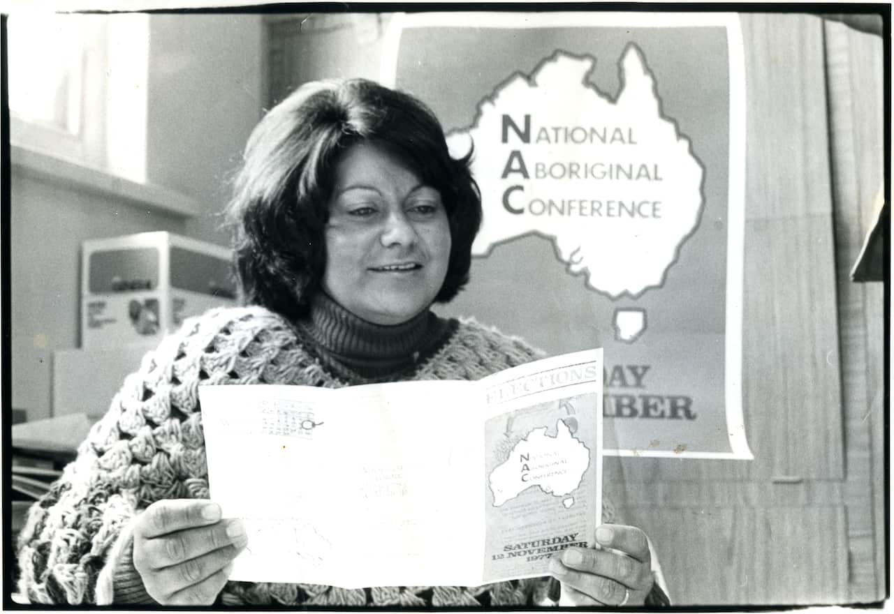 Black and white picture of a woman reading a flyer. There is a poster behind her featuring a map of Australia with the words National Aboriginal Conference written on it. 