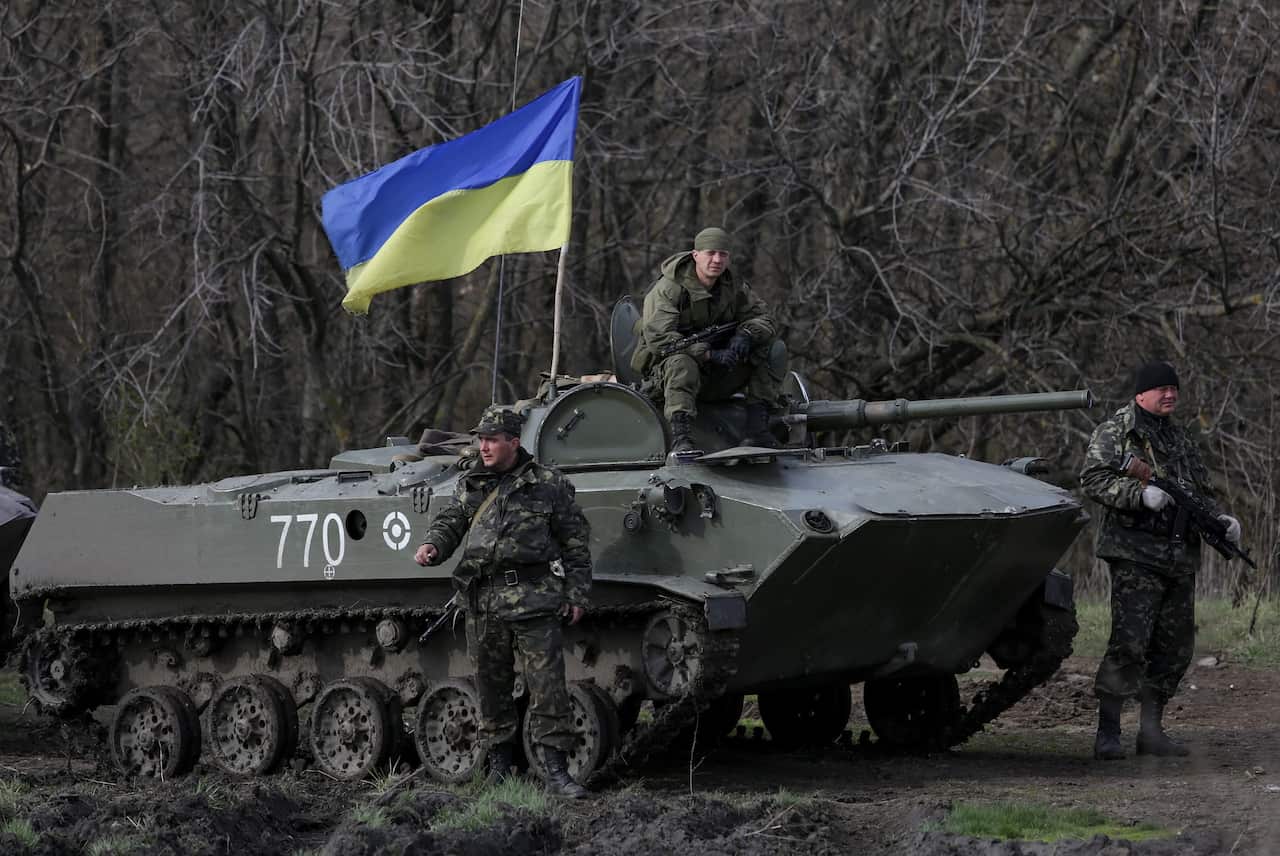 An army tank with a blue and yellow flag flying from it. A soldier is sitting on the tank, and there is a soldier standing on either side of it.