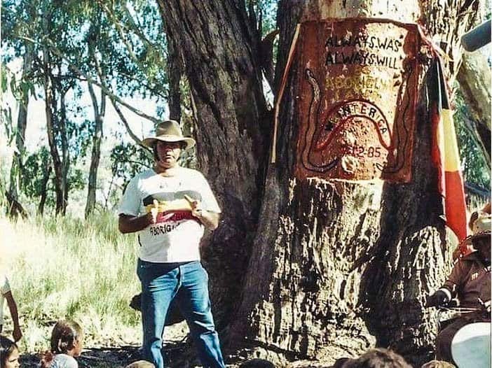 Mr Bates stands in front of a seated crowd. In the background is a tree with 'Always Was, Always Will Be' painted on it.