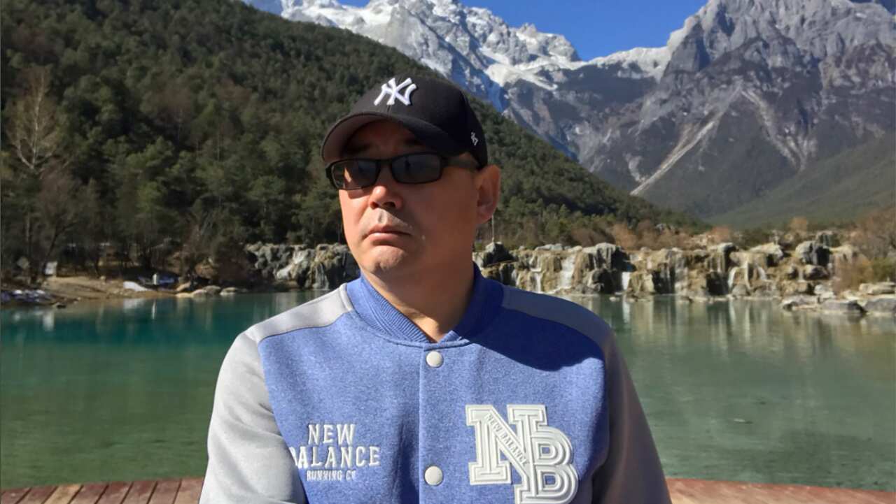 A man wearing a cap, sunglasses, and a bomber jacket standing outside.