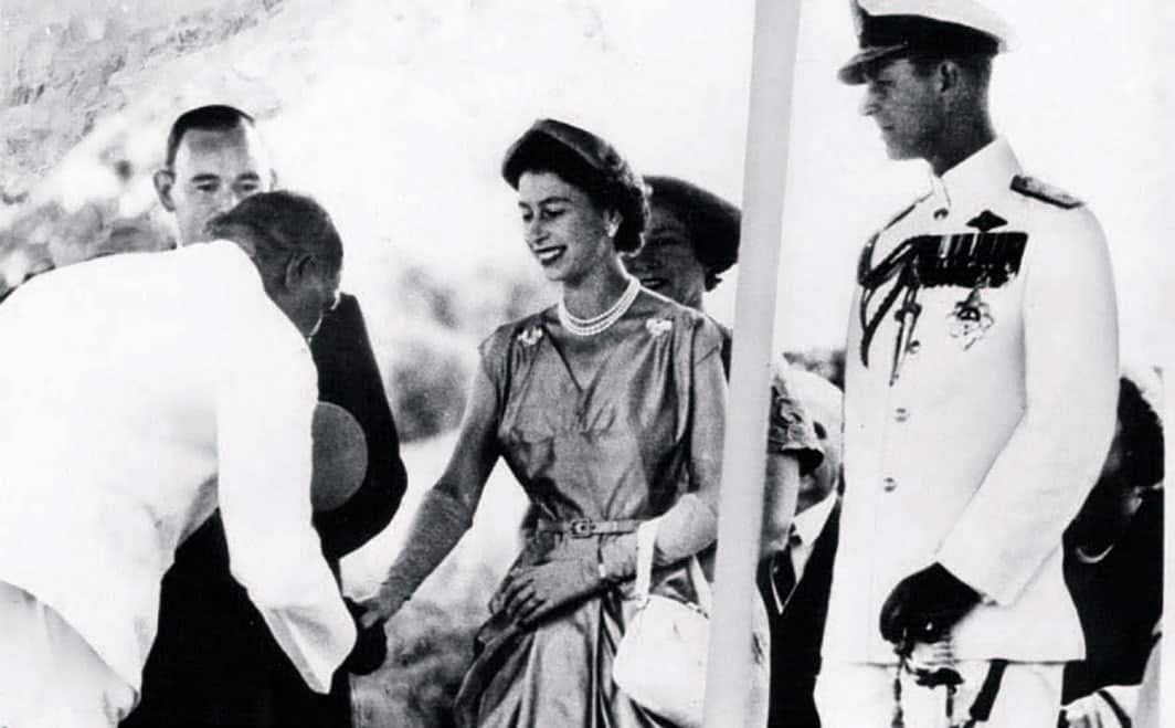 Black and white photo of Albert Namatjira bowing while shaking Queen Elizabeth's hand as Prince Phillip looks on. 