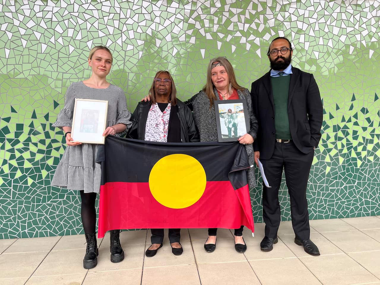 Three women and a man stand together and hold up an Aboriginal flag