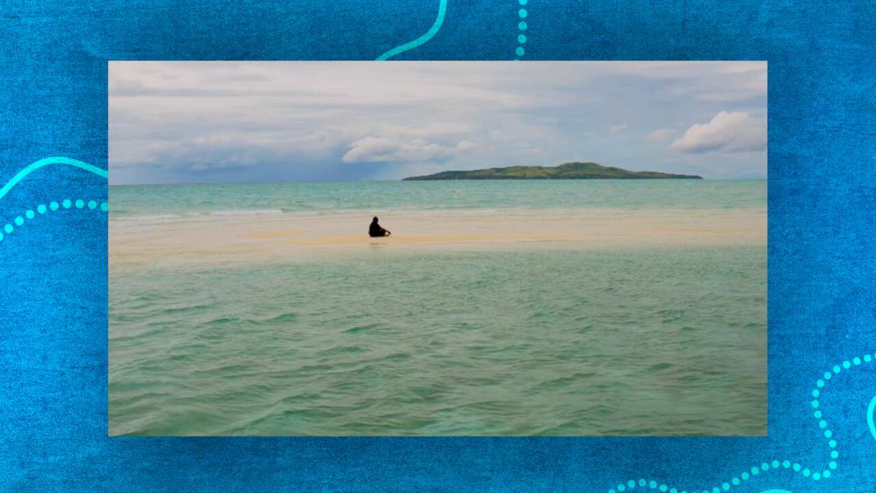 A still from a video of a person sitting in the distance cross-legged on a small patch of sand surrounded by sea water. There is an island in the distance. 