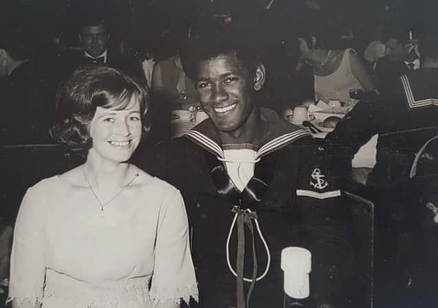 An Indigenous man wearing an Australian navy uniform and a white woman smile at the camera. They are sitting at a table amongst a large gathering in a hall.