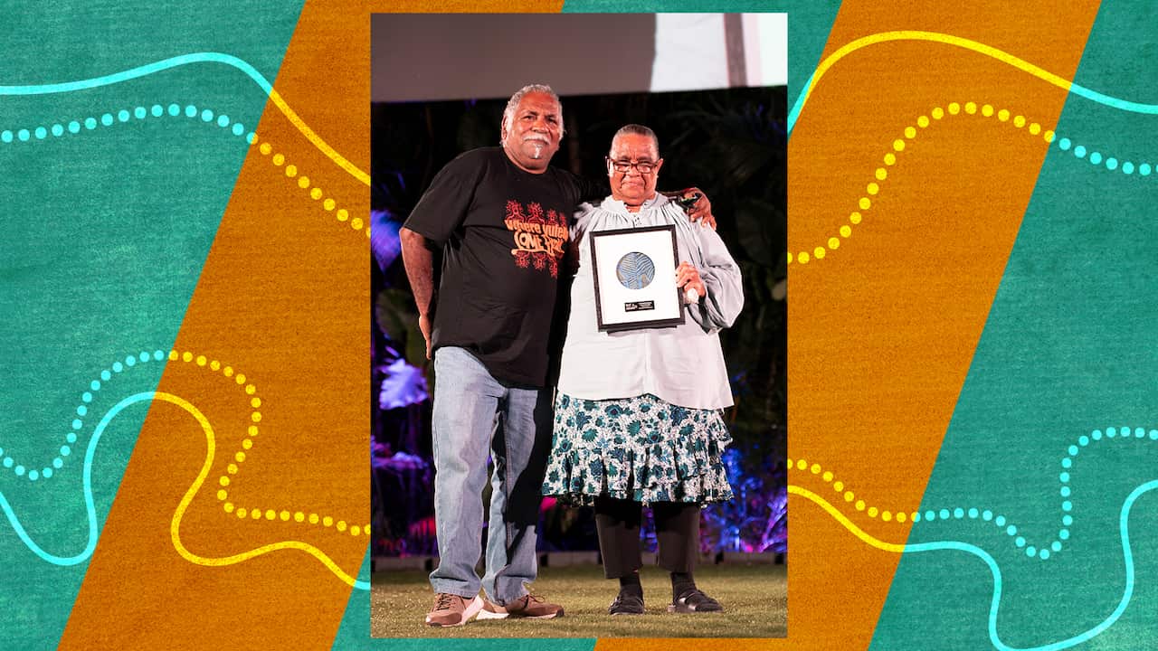 A middle aged couple, the bespectacled woman holding her award, with her casually dressed husband draping his arm around her shoulders.  