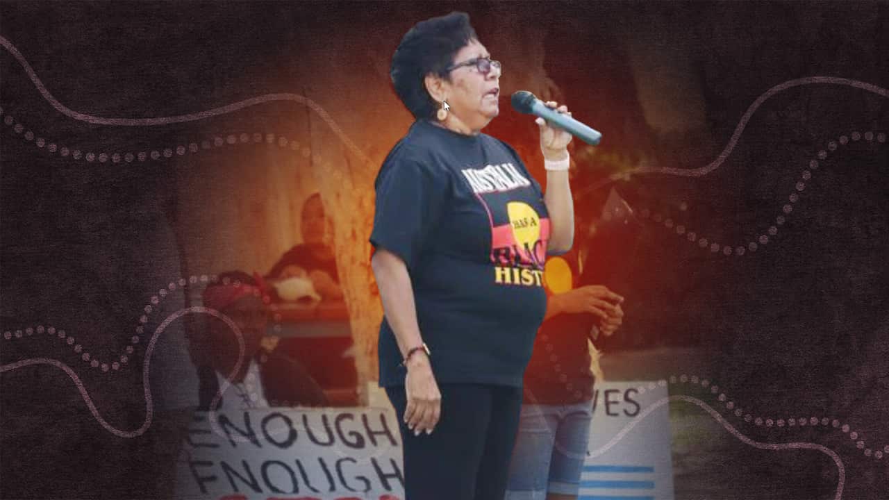 Florence Onus standing outside wearing an aboriginal flag tshirt speaks into a microphone, while people with protest signs sit behind her