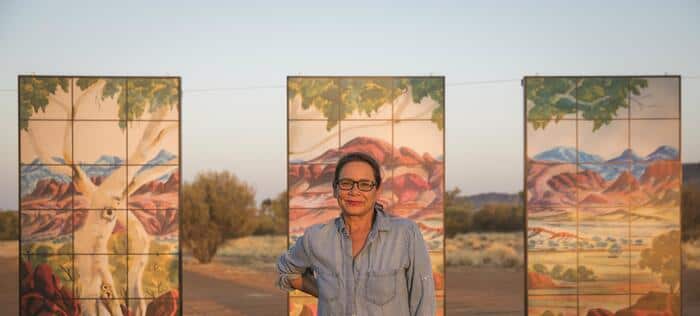 A woman standing in front of three panels of murals outside.