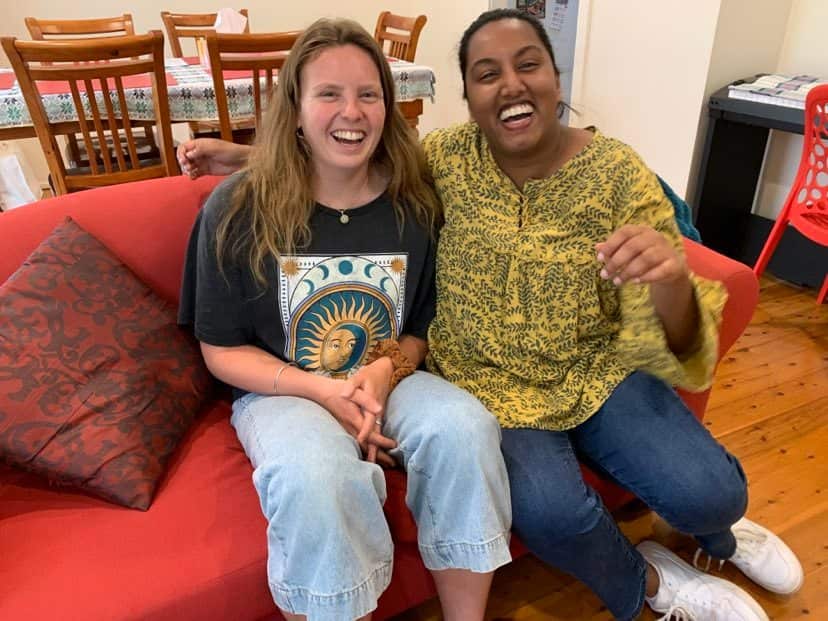 Kara (L) and Zoe (R) at Zoe's home, sitting on a red couch, laughing at the camera. 