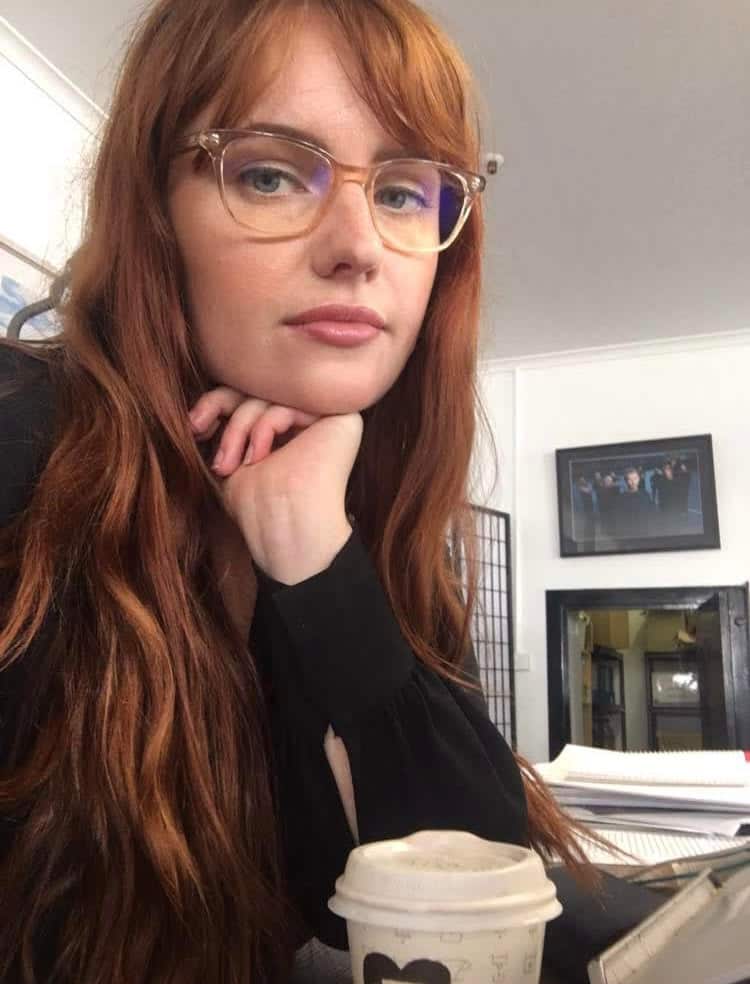 Laura Nagy in a black turtleneck, with long red hair, looking at the camera. There is a takeaway coffee on the table in front of her. 