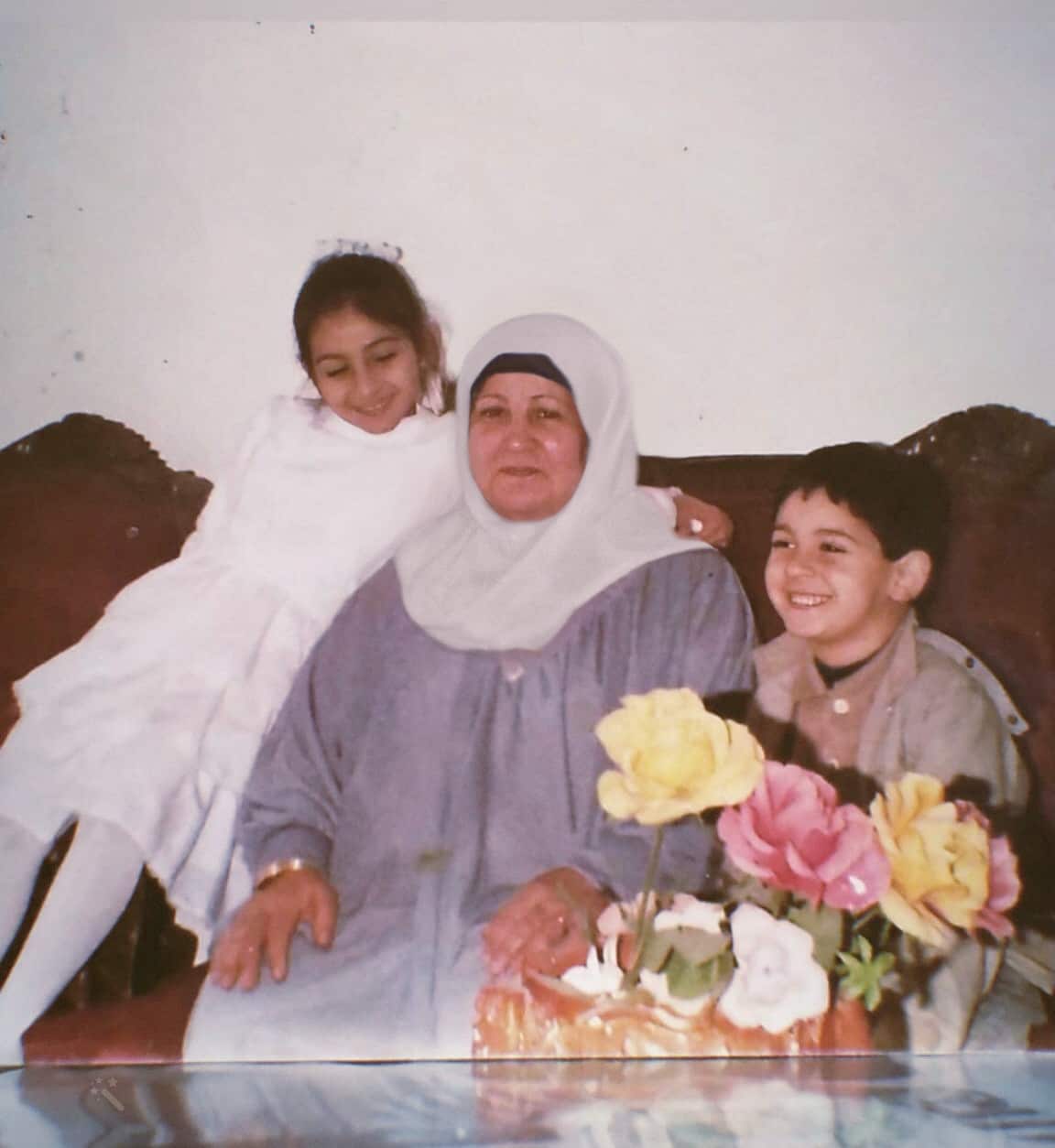 An older Muslim woman in headscarf with young children either side of her, all sitting on a couch.