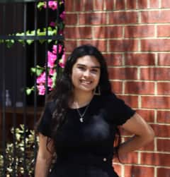 Young woman of Peruvian descent, standing in front of brick wall and smiling.